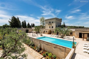 A pool area with a clear blue pool, surrounded by a stone wall and various plants. There are lounge chairs and a building with a tower-like structure in the background.