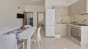 A compact kitchen with a refrigerator, stove, and sink. There is a dining area with a table and chairs. - 1064 PMR - Appartamento Dei Delfini (Porto Cesareo)