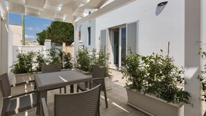 A covered patio with a table and chairs, surrounded by potted plants and a white wall. - 1064 PMR - Appartamento Dei Delfini (Porto Cesareo)
