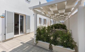 A covered outdoor area with a pergola, potted plants, and a view of a building with windows and shutters.