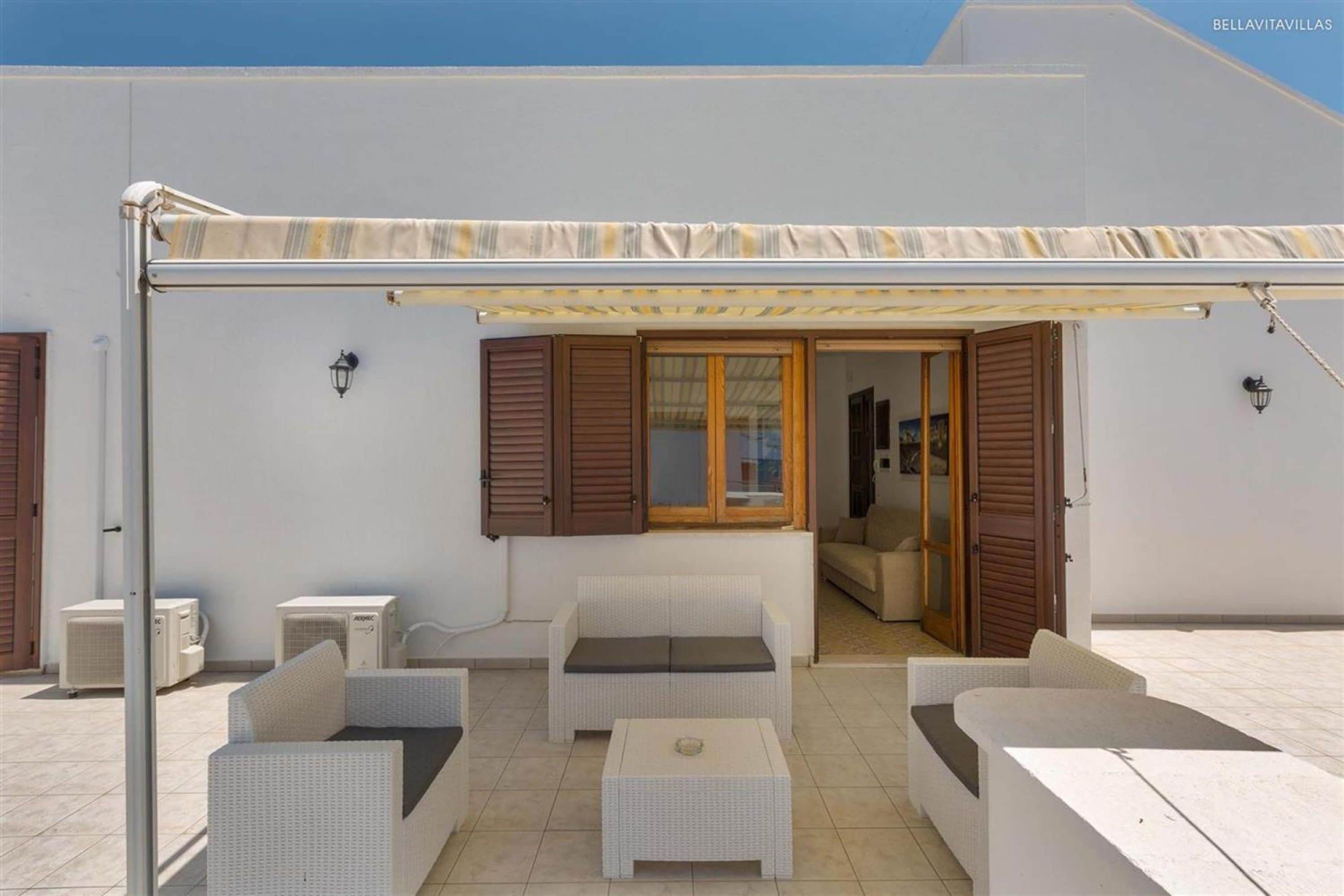 A covered outdoor seating area with white wicker furniture, including sofas and a coffee table, under a striped awning.