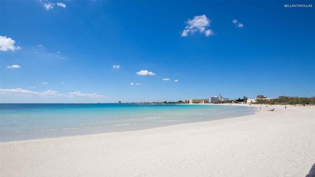 Ein Strand mit weißem Sand und klarem, blauem Wasser. Im Hintergrund sind Gebäude zu sehen, im Vordergrund ein Holzzaun.