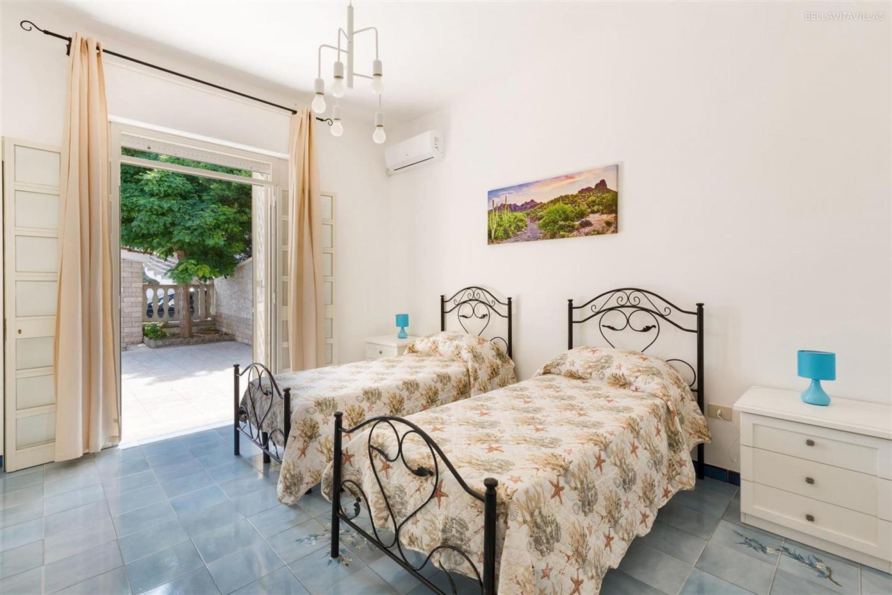 A bedroom with two beds, a white dresser, a painting, and an open door leading to a courtyard.