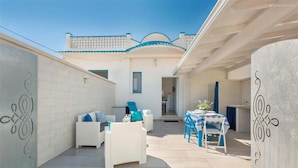 Un amplio patio exterior con mobiliario de mimbre blanco, una mesa de comedor con mantel a cuadros azul y blanco, y una zona de cocina visible a través de un vano abierto.