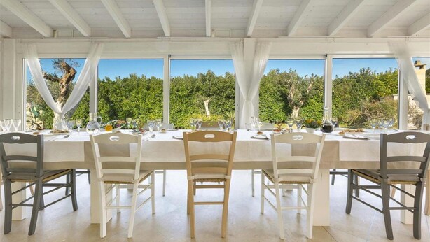 A dining area with a white table set for six, surrounded by chairs, large windows, and a view of greenery. - 1738 Tenuta Degli Ulivi (Melendugno)