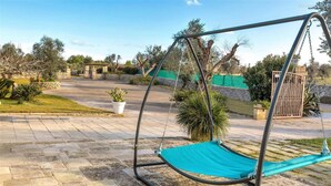 A hammock swing in a courtyard with stone paving, a gate, and surrounding greenery. - 1738 Tenuta Degli Ulivi (Melendugno)