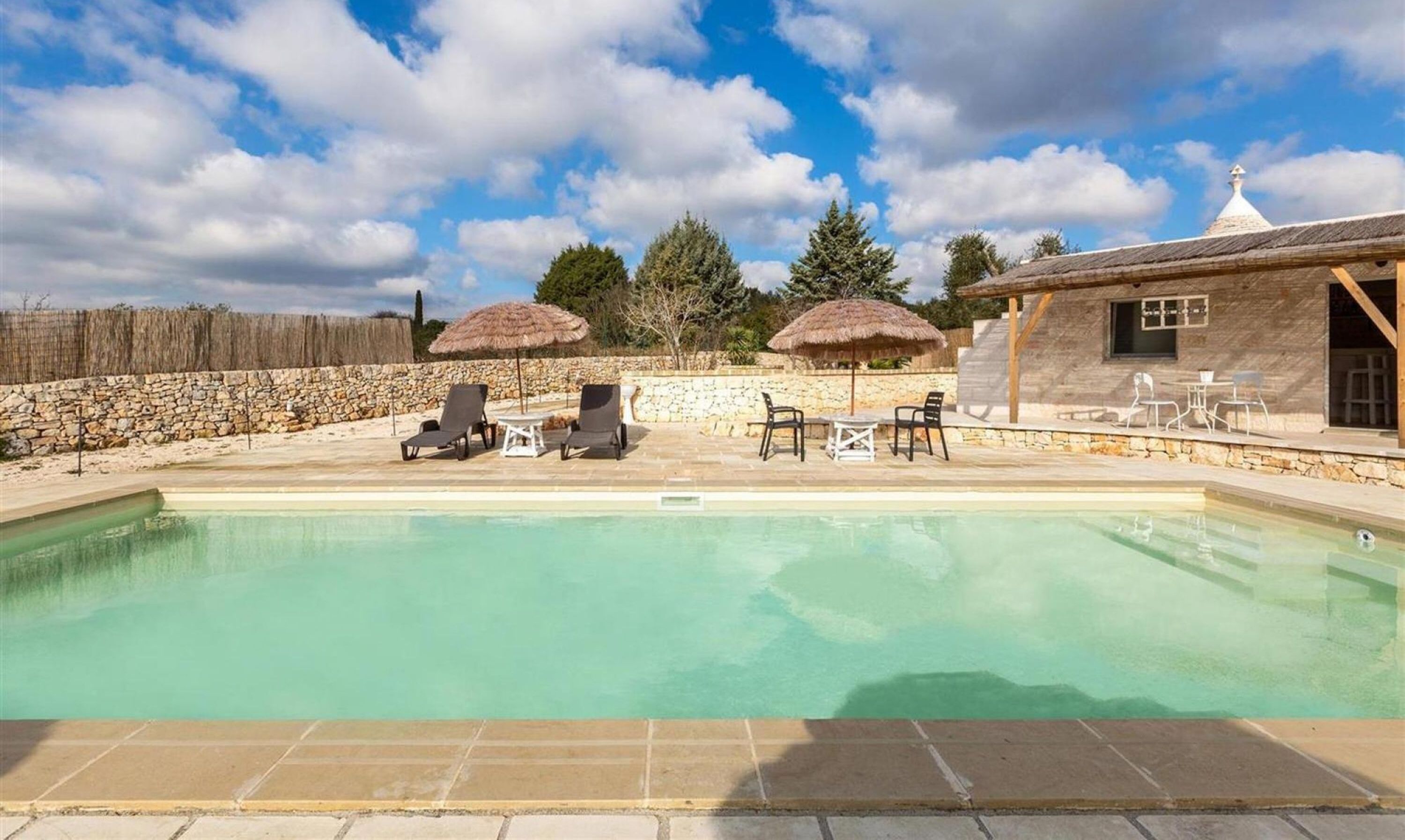 A swimming pool with lounge chairs and umbrellas, surrounded by stone walls and a stone building with a tiled roof.