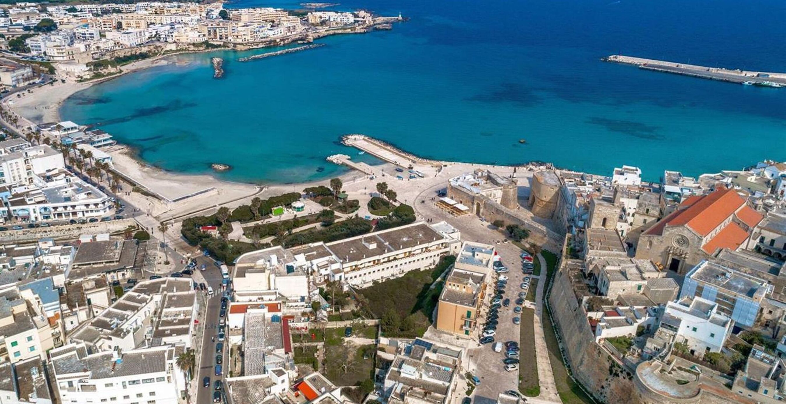 An aerial view of a coastal town with a mix of residential and commercial buildings, a prominent fort-like structure, and a clear blue sea.