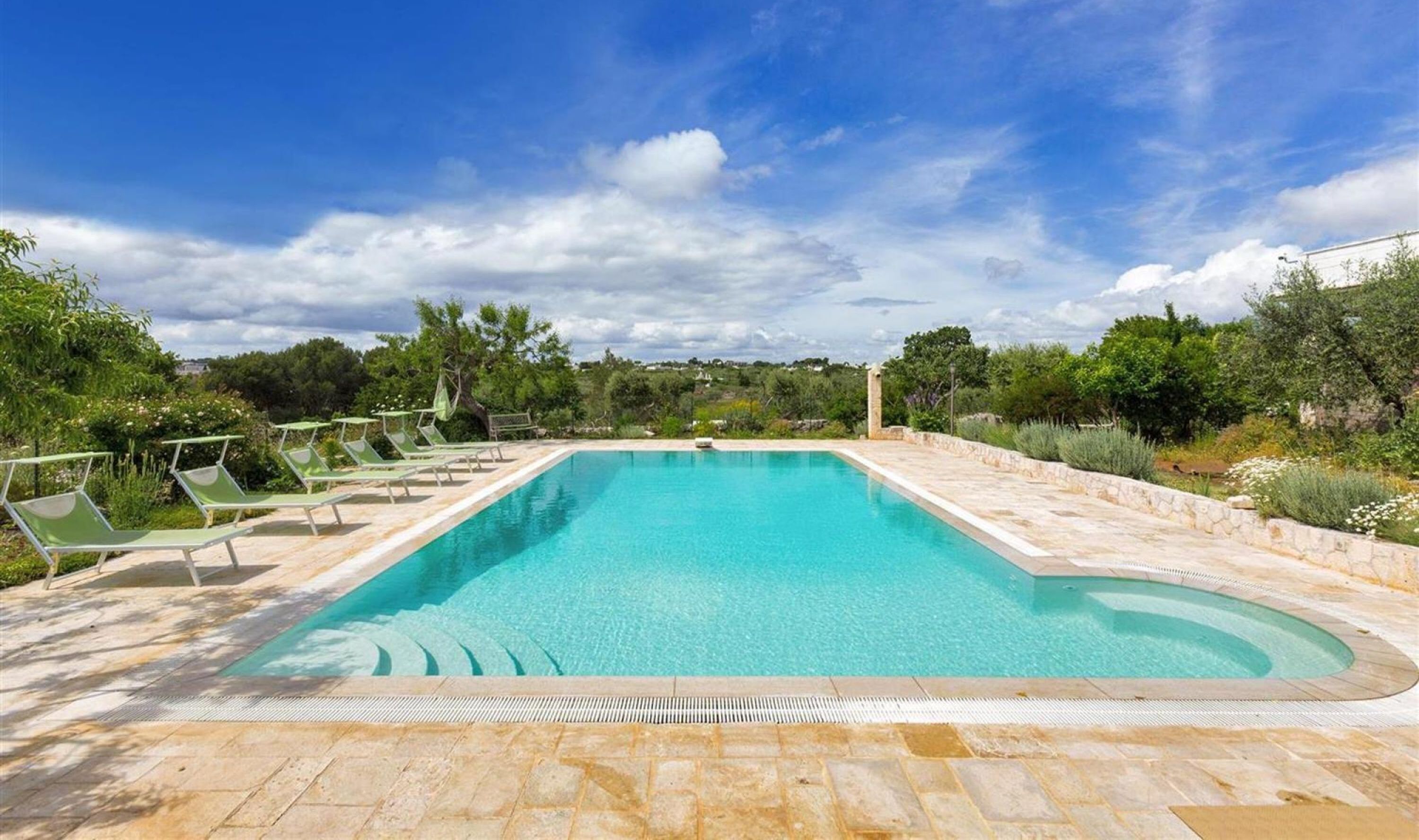 A rectangular pool with a stone border, surrounded by greenery and a clear sky.