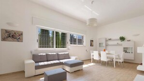 En rummelig opholdsstue med en hvid sofa, et spisebord med stole, og store vinduer der giver naturligt lys.