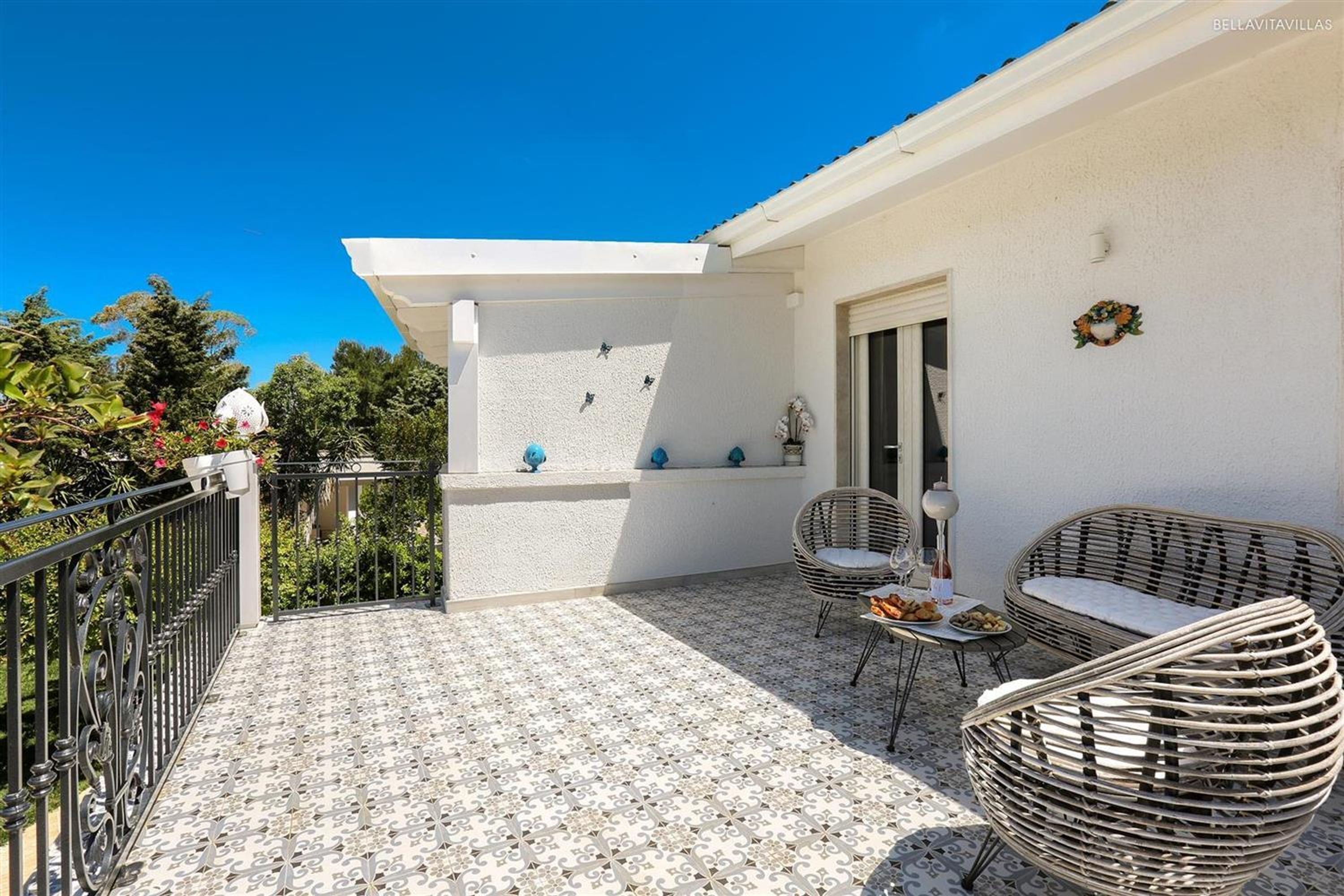 Un balcon doté d’un sol à motifs, de meubles en osier et d’un mur blanc avec une porte et une fenêtre.