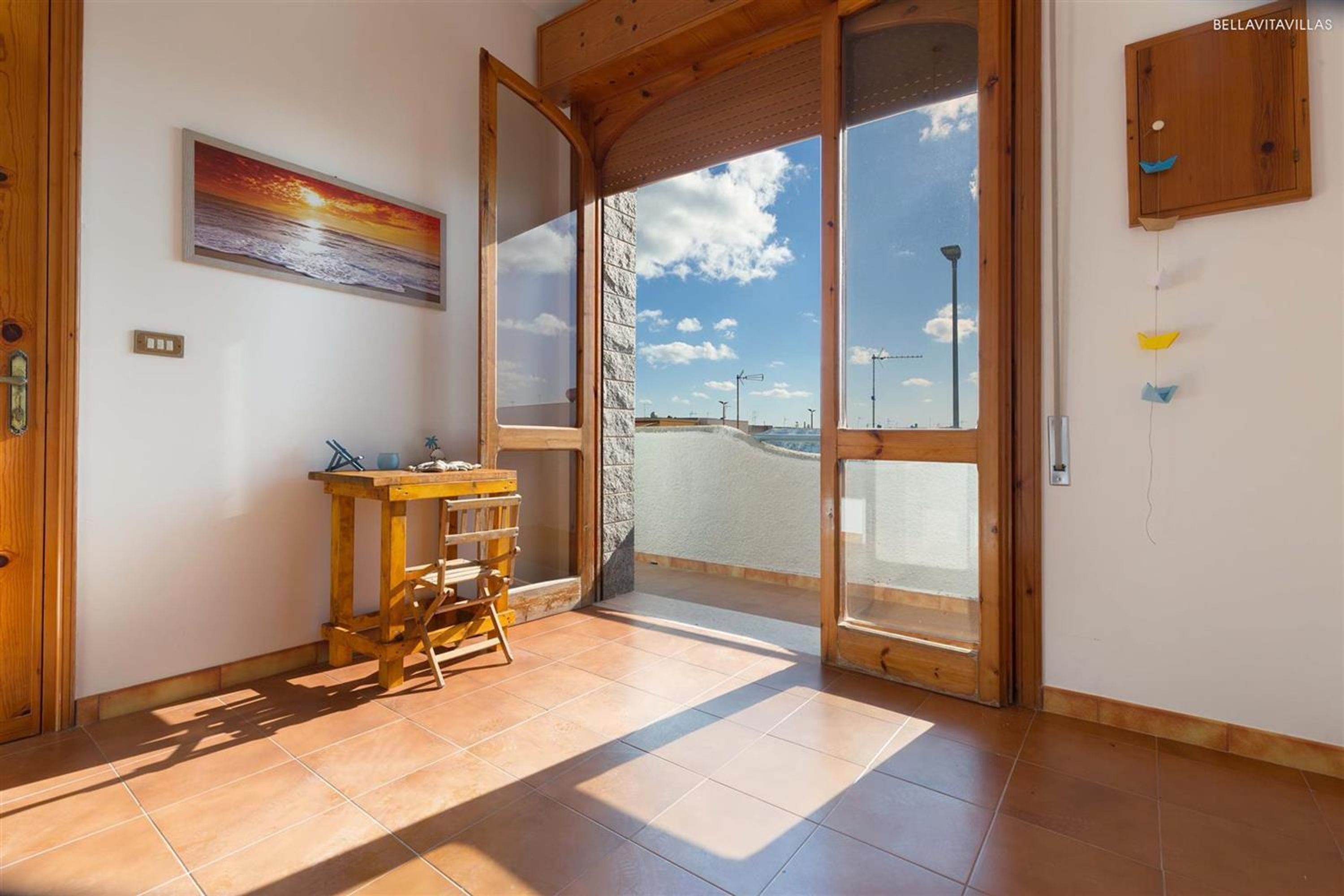 A room with a wooden table, a chair, a painting of a sunset, and a large glass door leading to a balcony with a view of the sky and clouds.