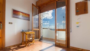 A room with a wooden table, a chair, a painting of a sunset, and a large glass door leading to a balcony with a view of the sky and clouds. - 2191 Appartamento Blue Dreams by Barbarhouse (Melendugno)