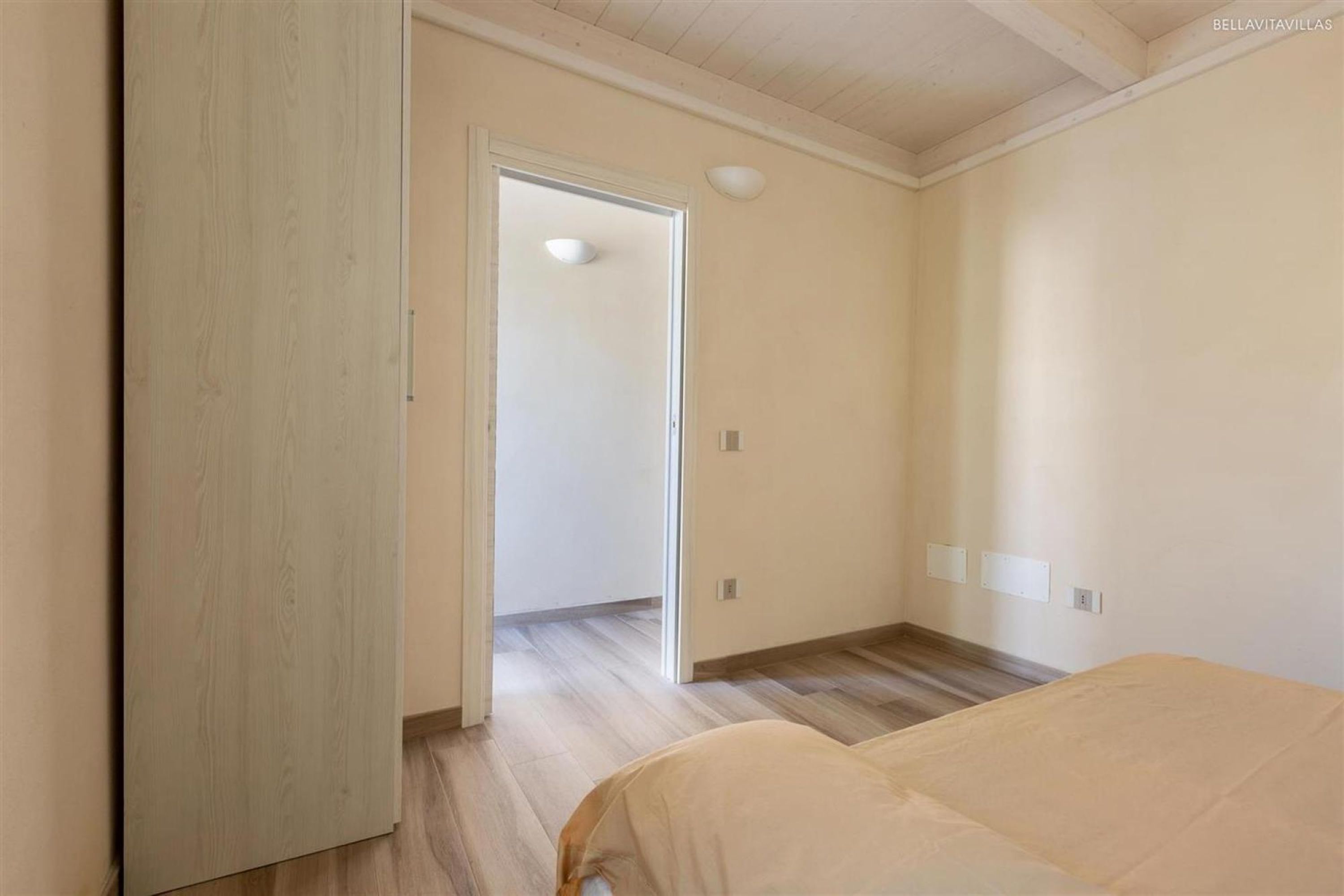 A bedroom with a wooden wardrobe, a bed, and a door leading to another room.