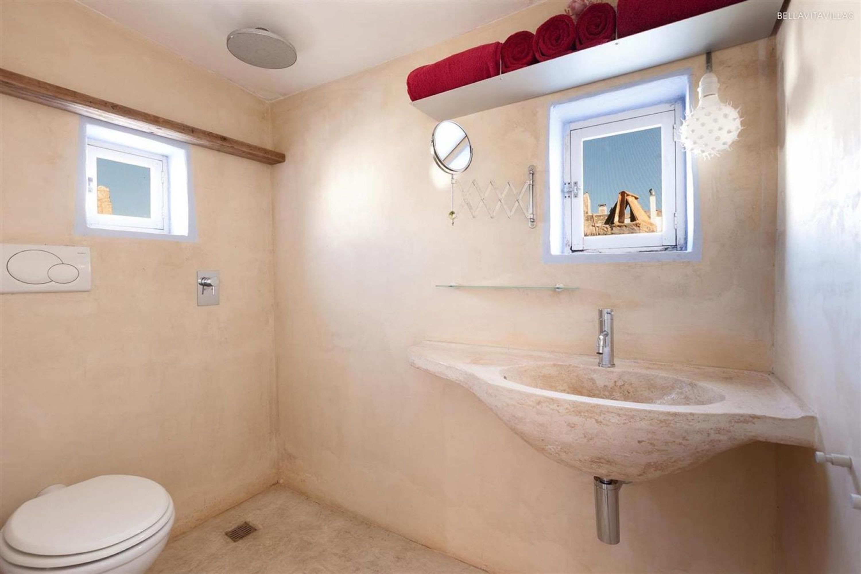 A bathroom with a white toilet, a beige sink, a window with a mirror, and a towel rack with red towels.