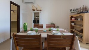 Un coin repas avec une table dressée pour quatre personnes, entourée de chaises en bois. On y trouve une bibliothèque avec des objets décoratifs et un petit modèle de bateau posé sur le dessus.