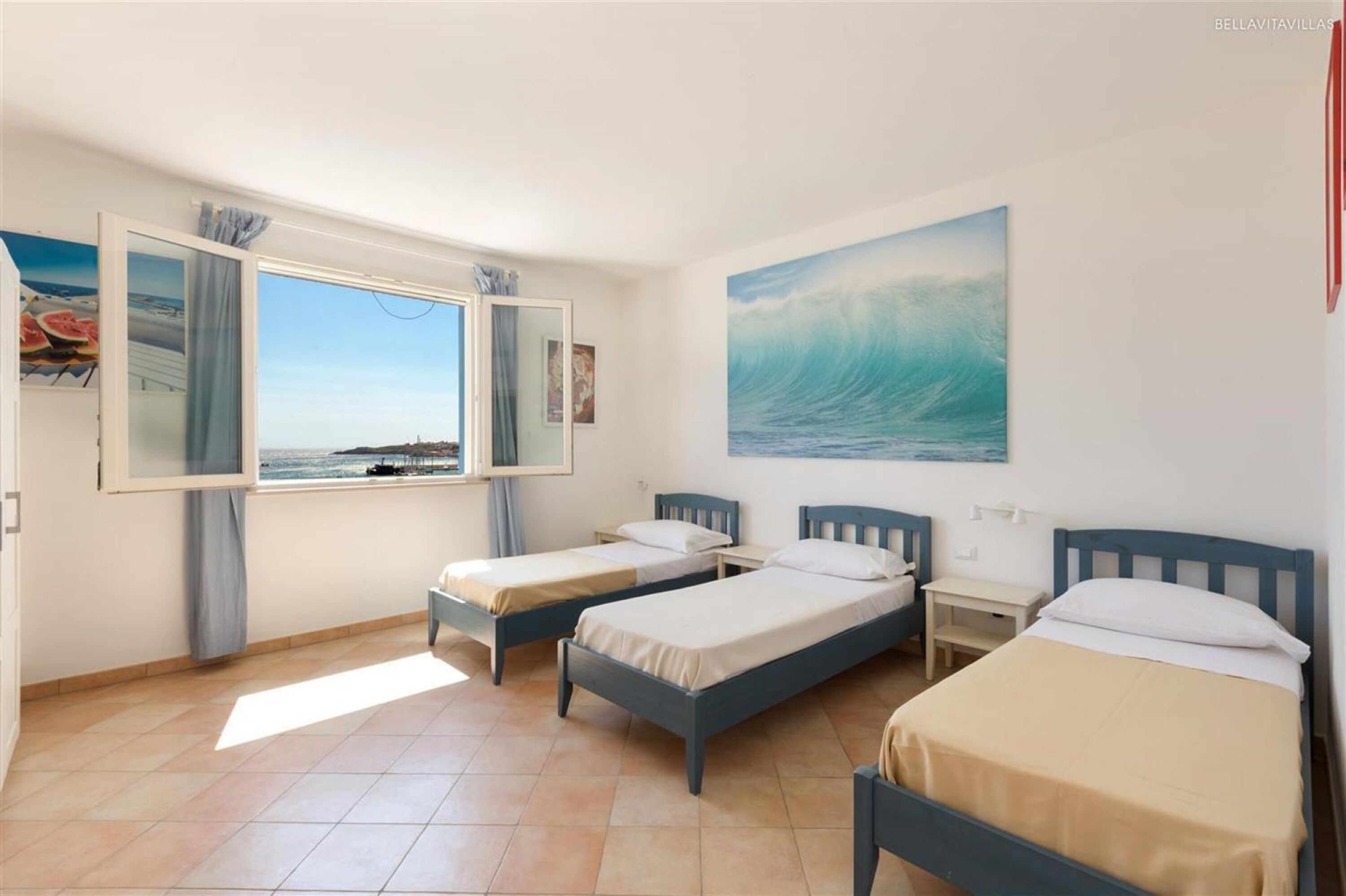 A room with two single beds, a large window with a view of the sea, and a painting of a wave on the wall.
