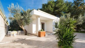 A small, white building with a tiled patio, a table with chairs, and a large plant. - 1368 Villetta La Pineta 1 (Porto Cesareo)