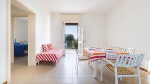 A bright room with a dining table set for four, a bed with striped bedding, and a view through an open door to a balcony. - 1368 Villetta La Pineta 1 (Porto Cesareo)