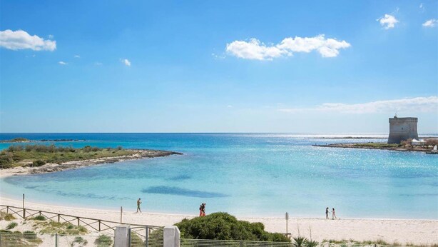 A beach with clear water, a distant tower, and a fenced area.