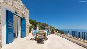 A terrace with a table and chairs, a stone building with blue shutters, and a view of the sea.