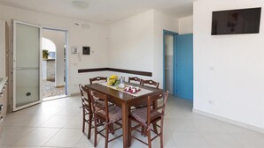 A dining area with a wooden table and chairs, a TV mounted on the wall, and a door leading to an outdoor area. - 1515 Villetta Raffaele (Nardò)