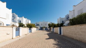 A paved pathway leading to a row of white buildings with blue doors and windows, surrounded by low stone walls and greenery. - 1515 Villetta Raffaele (Nardò)