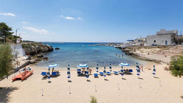 Egy tiszta, kék vízű strand, sziklás parttal és egy fehér homlokzatú épülettel.