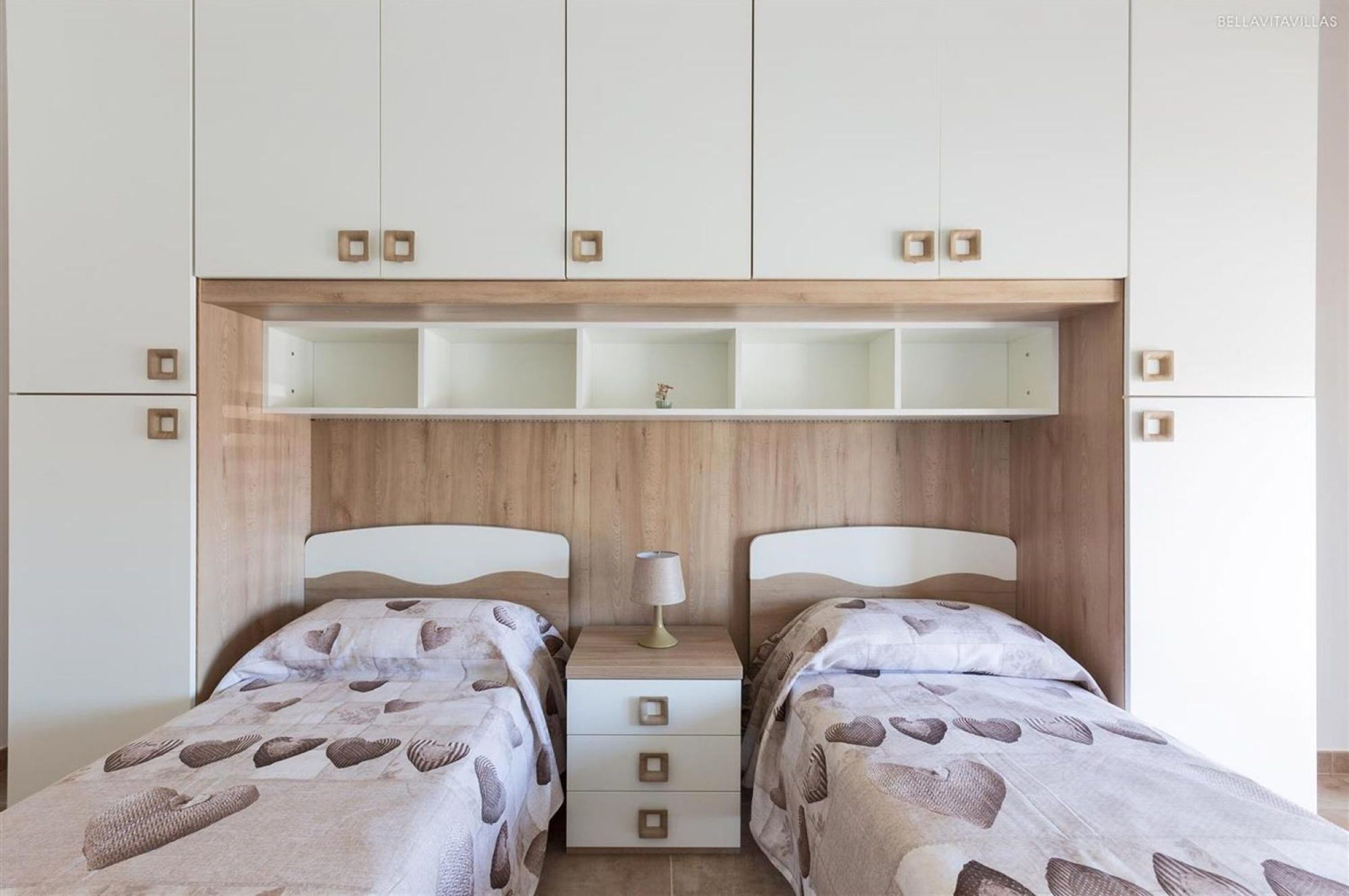 A bedroom with two beds, a wooden headboard, a nightstand, and a shelf above the beds.