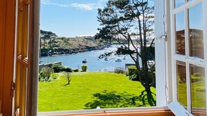 Une vue depuis une pièce dotée d’une grande fenêtre, donnant sur une zone verdoyante et un plan d’eau où se trouvent des bateaux.