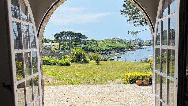 Une porte ouverte donne sur un patio dallé de pierres, d’où l’on peut admirer un paysage côtier comprenant une étendue d’eau et de la végétation.