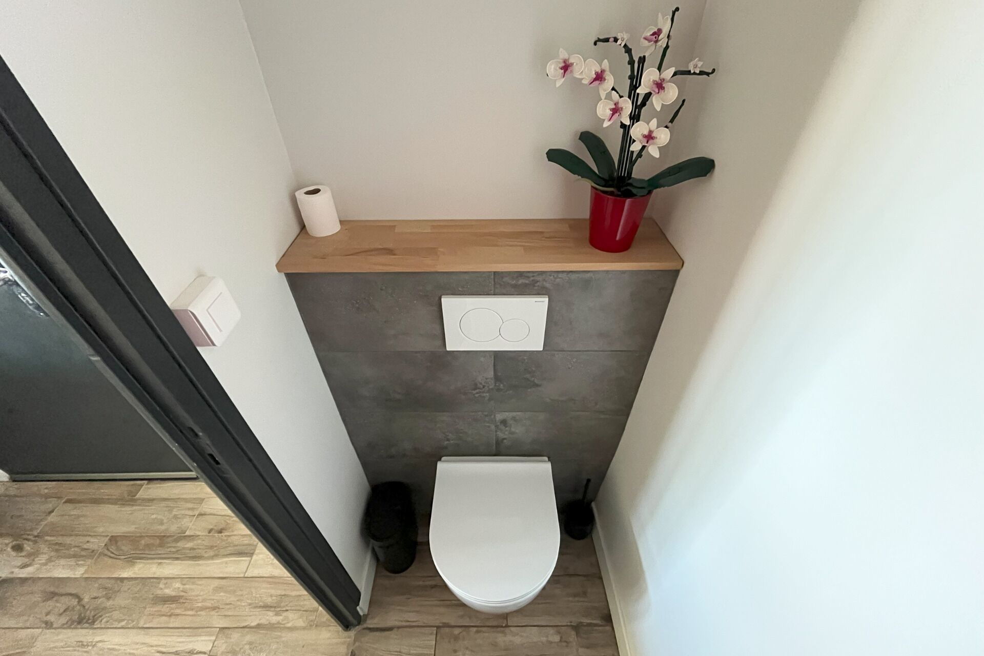 A small bathroom with a toilet, a wooden shelf with a toilet paper roll, a potted orchid, and a wall-mounted light switch.