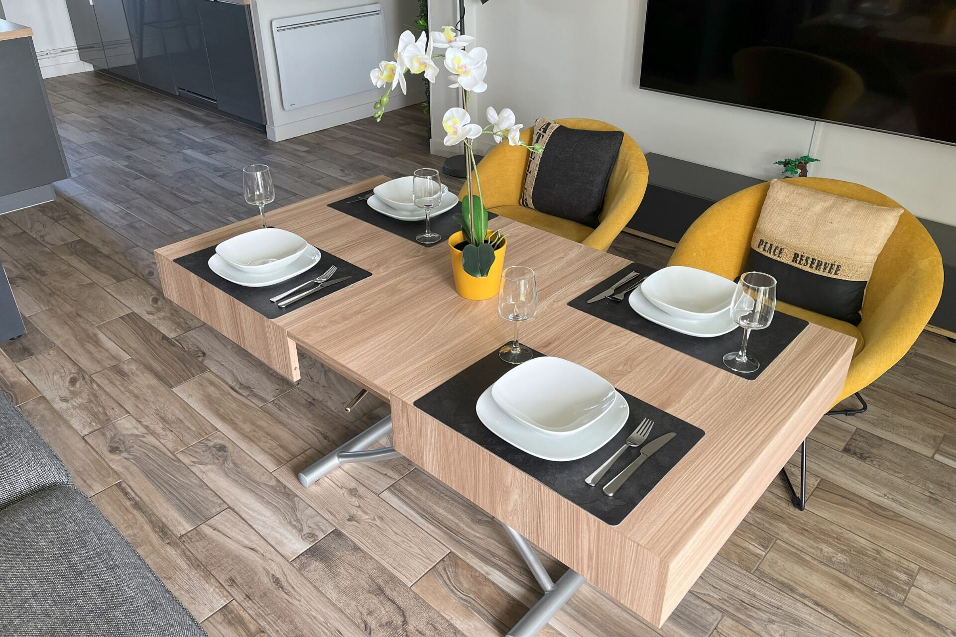 Un coin repas avec une table en bois dressée pour quatre personnes, agrémentée de sets de table noirs, d’assiettes blanches et de couverts.