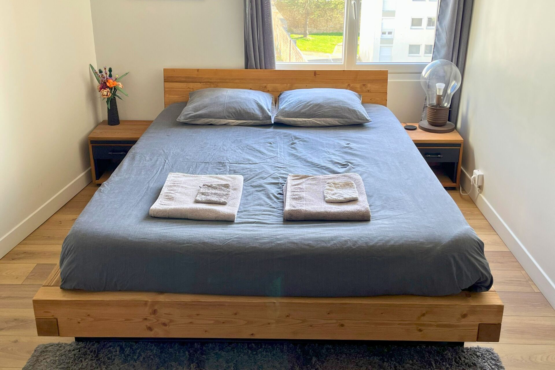 Un lit bien fait, avec deux serviettes, une tête de lit en bois, une table de chevet avec une lampe et une fenêtre donnant sur des immeubles.