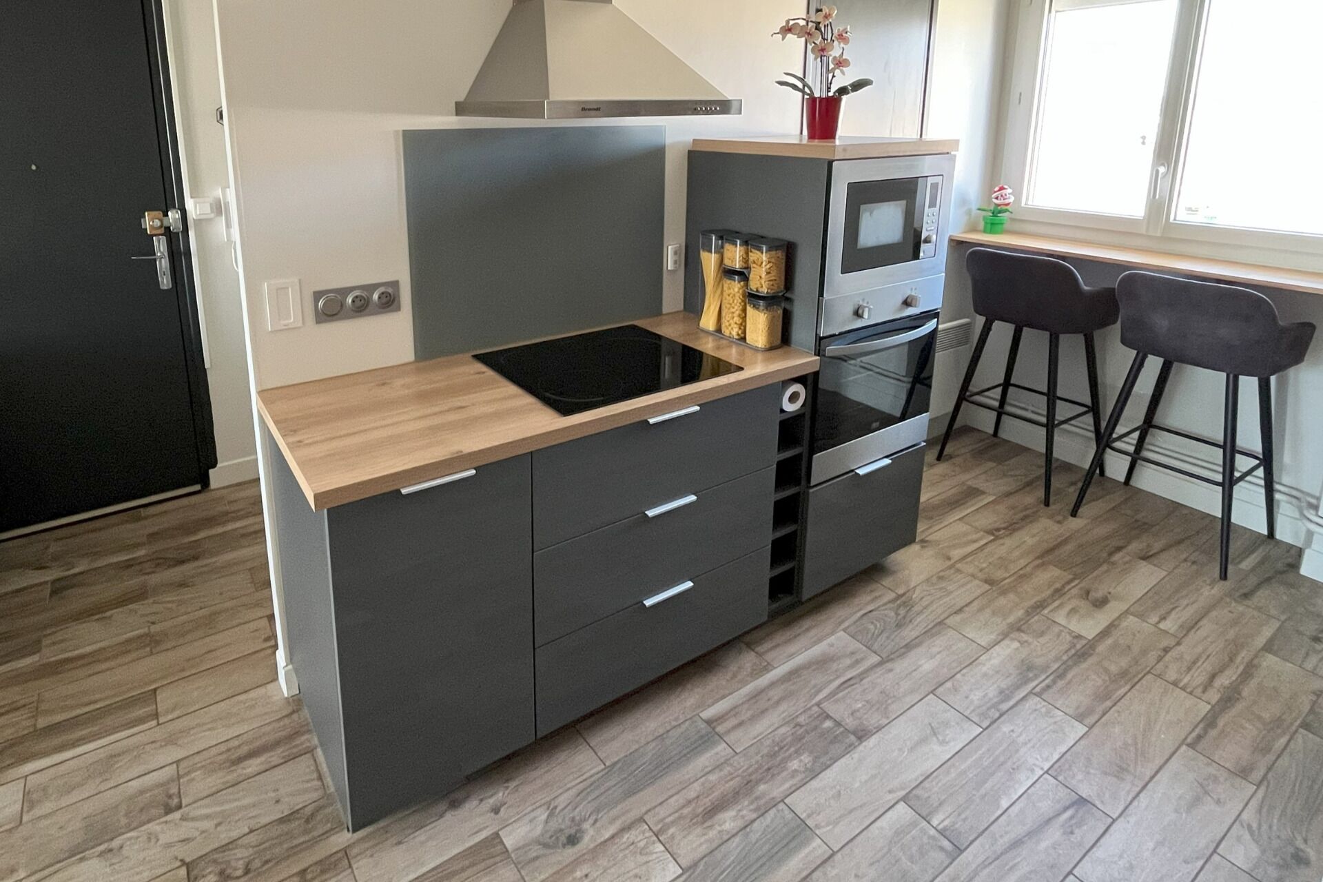 A modern kitchen with a black refrigerator, a built-in oven, a microwave, and a wooden countertop. There are two black bar stools and a window with a view of the outside.