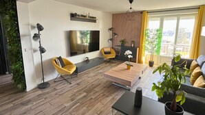 A modern living room with a flat-screen TV, a yellow armchair, a wooden coffee table, and a large window with curtains.