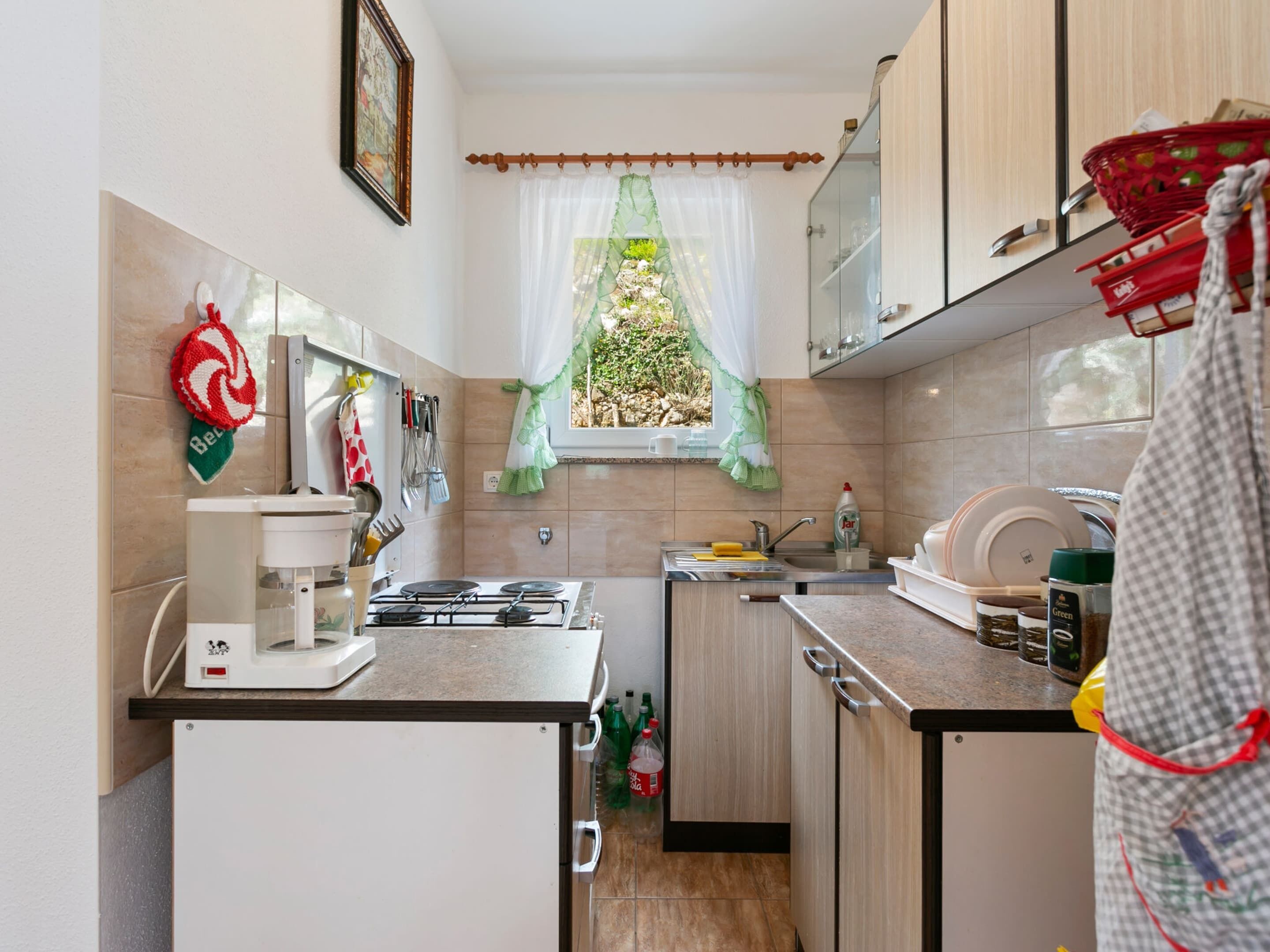 Une cuisine compacte équipée d’une cuisinière à gaz, d’un évier et de meubles en bois. Une fenêtre avec un rideau vert est visible, et divers ustensiles de cuisine sont accrochés au mur.