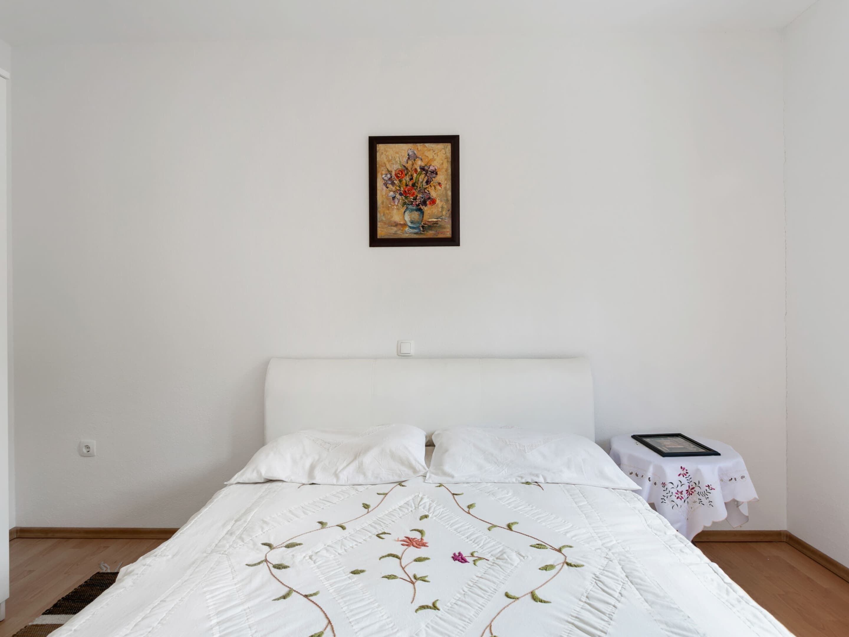Une chambre à coucher avec un lit, du linge de lit blanc, une table de chevet avec un petit plateau, et une œuvre d’art encadrée accrochée au mur.