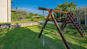 Un ensemble de balançoires en bois, installé dans une cour verdoyante, avec un mur de pierres et un bâtiment en arrière-plan.