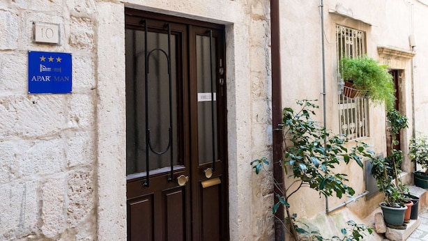 A building with a dark brown door, a blue sign with stars and the word "APARTMAN", and potted plants on the right side. - Apartment Neve - Two Bedroom Apartment (Dubrovnik)