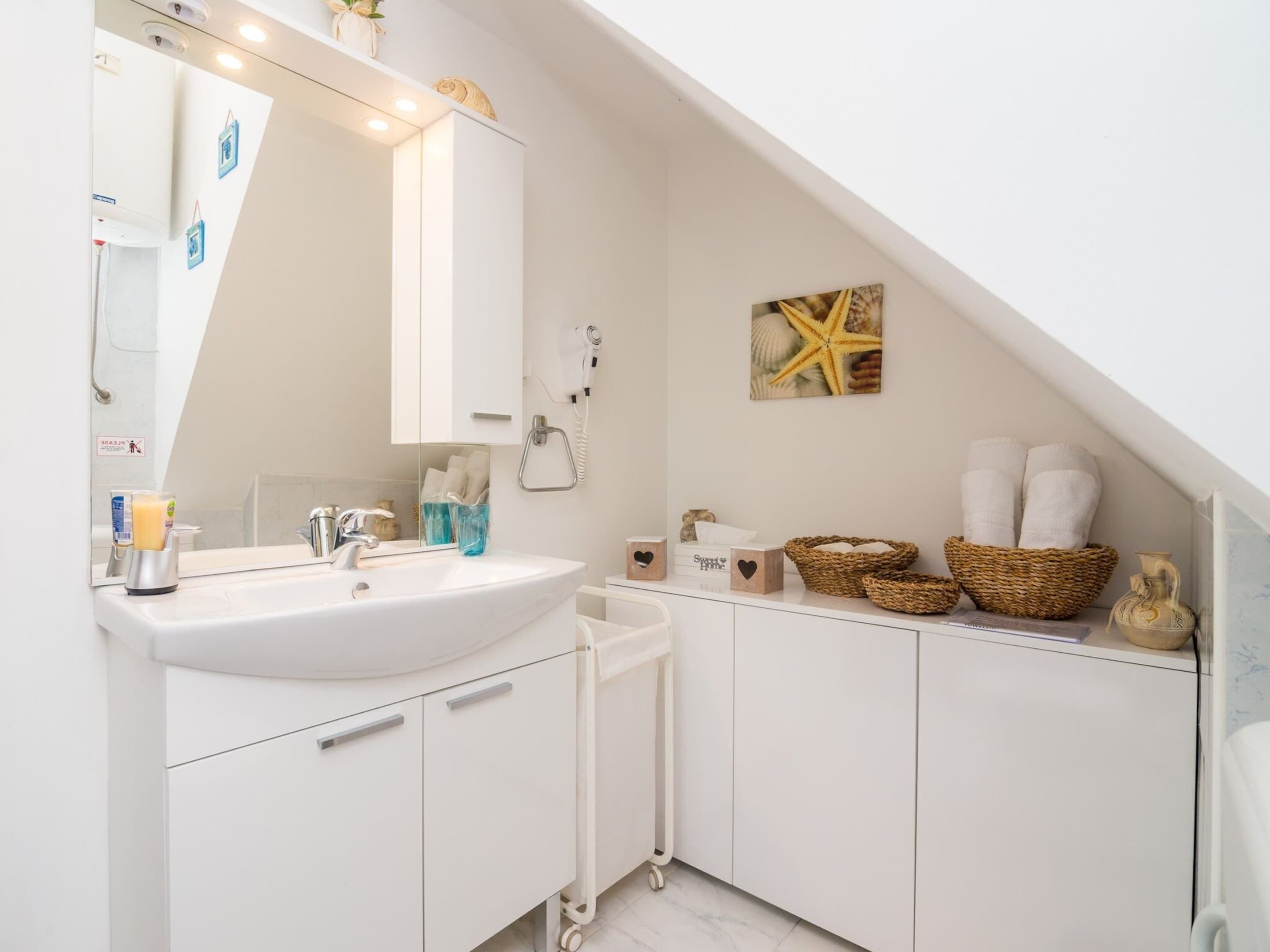 Een moderne badkamer met een witte wastafel, een badkamermeubel en een grote spiegel. Op het meubel staan manden en handdoeken, en aan de muur een decoratieve zeester.