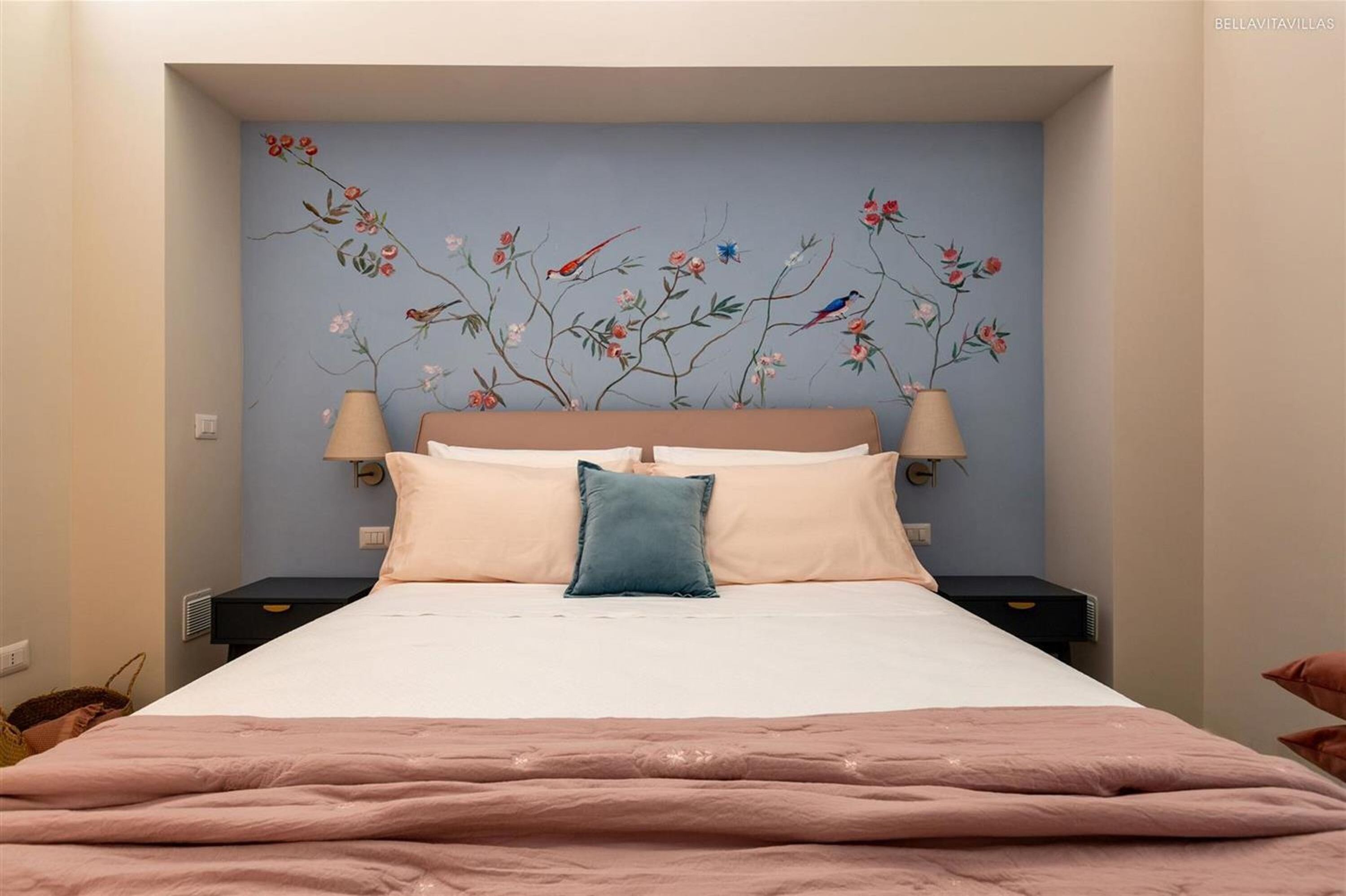A bedroom with a bed, two bedside tables, a nightstand, a wall-mounted lamp, and a decorative floral mural on the wall.