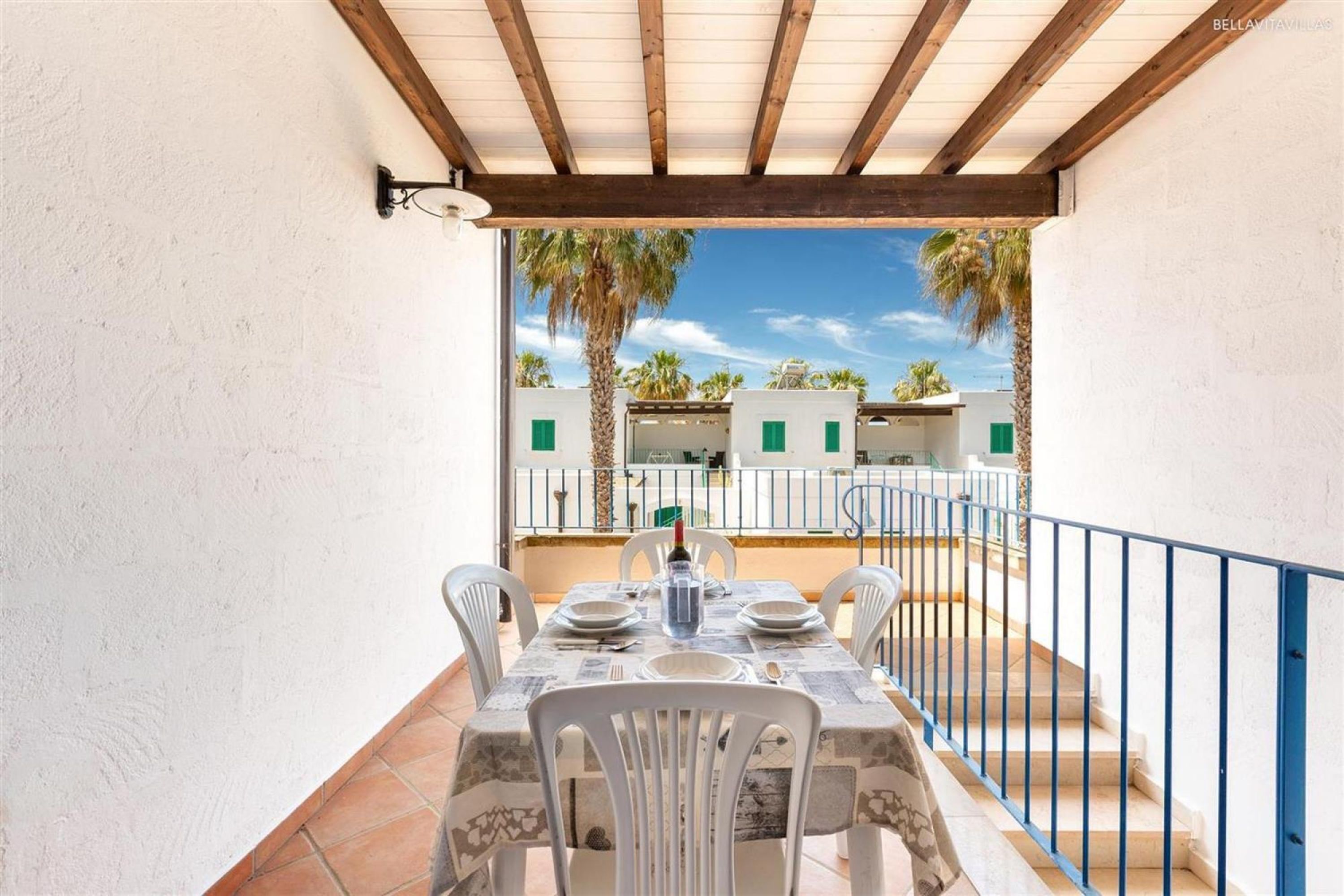 A dining area with a table set for four, white chairs, and a view of a courtyard with palm trees and buildings.