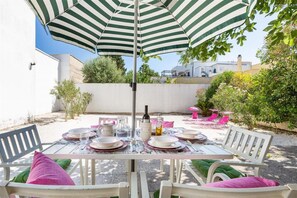 Un área de comedor al aire libre con una mesa preparada para cuatro personas, rodeada de sillas blancas con cojines verdes, bajo un paraguas a rayas.
