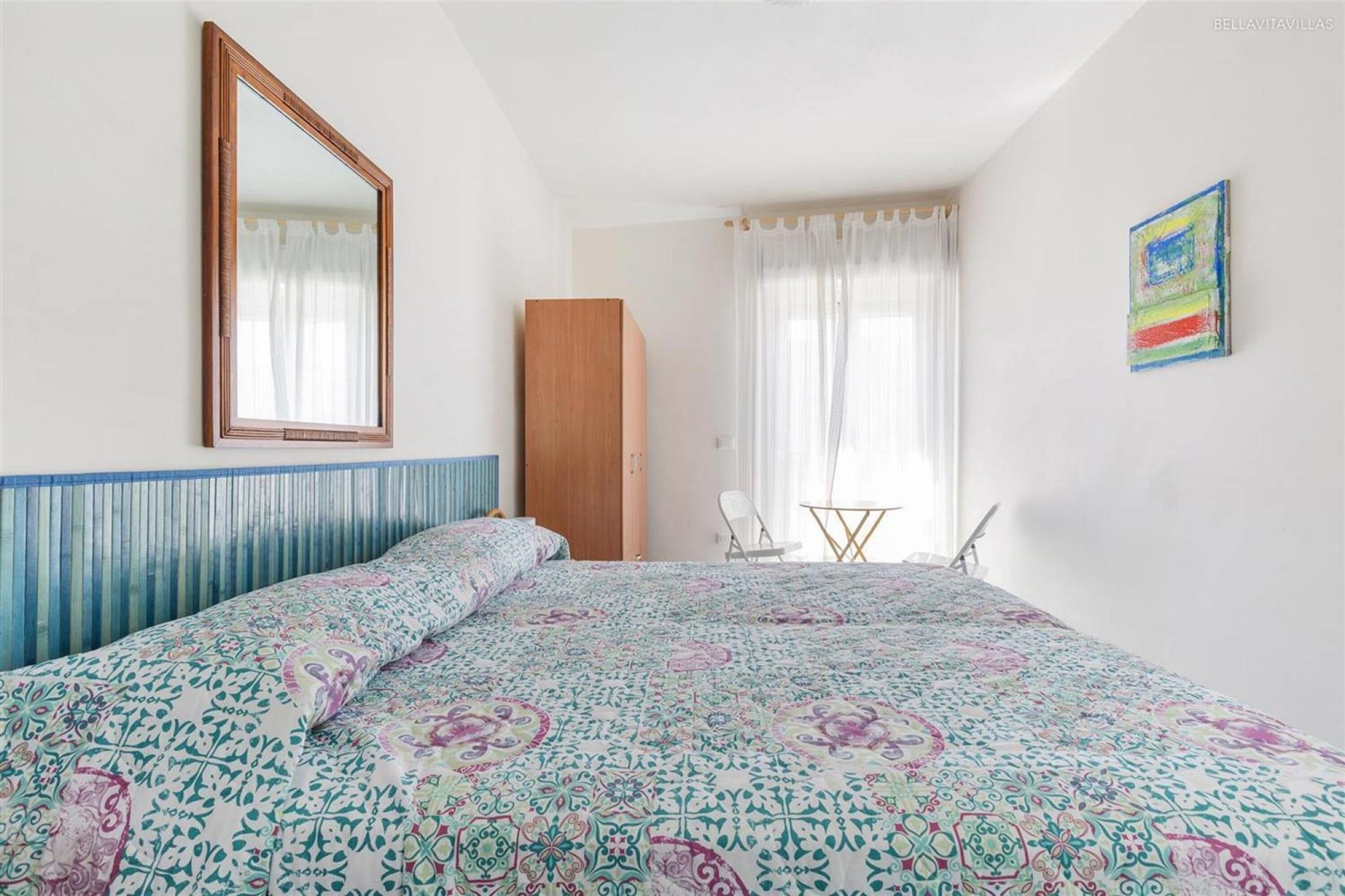 Ein Schlafzimmer mit einem Bett, einem Holzschrank, einem kleinen Tisch mit Stuhl und einem farbenfrohen Bild an der Wand.