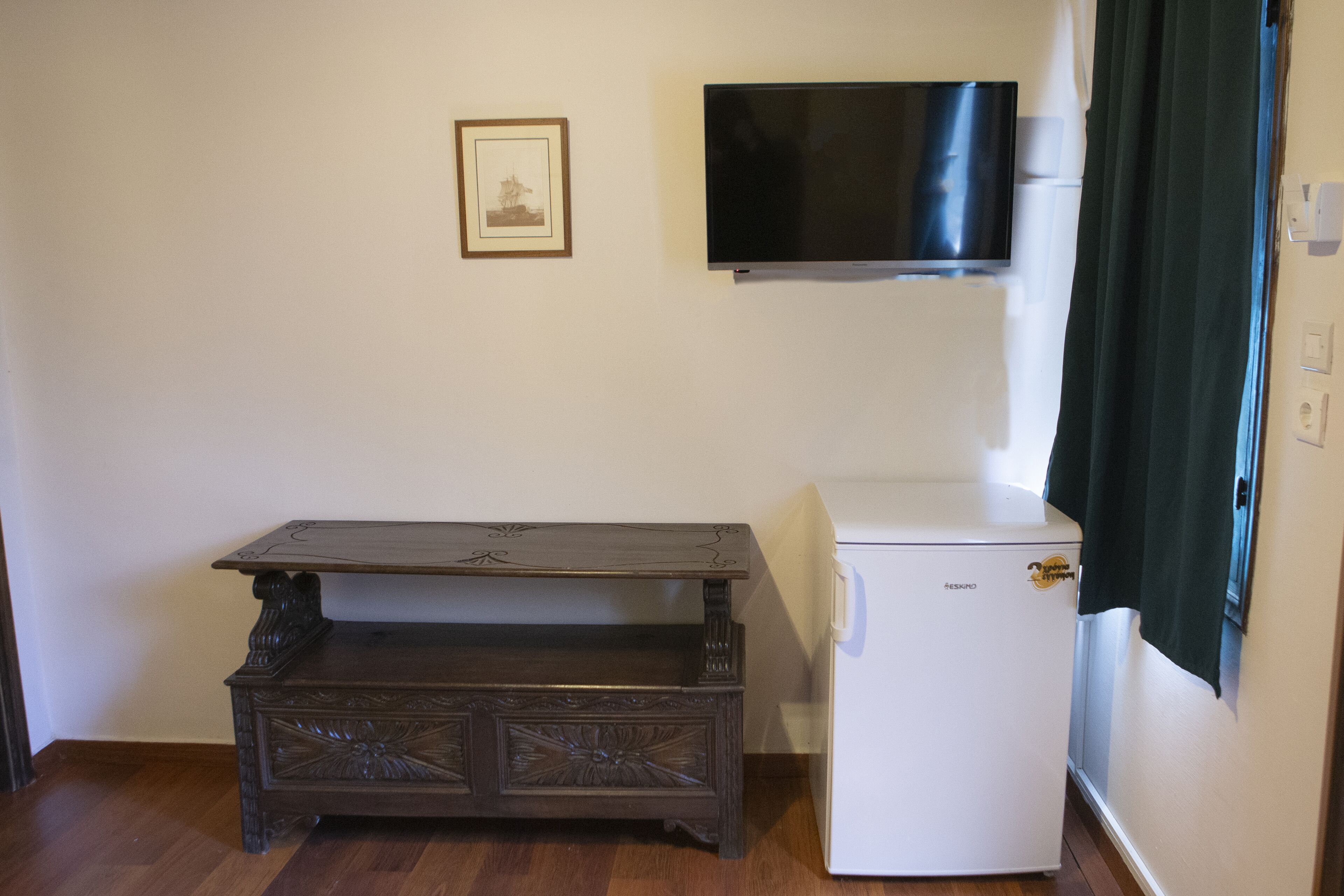 Una habitación pequeña con una mesa de madera, un refrigerador blanco, un cuadro en la pared y un televisor de pantalla plana sobre la mesa.