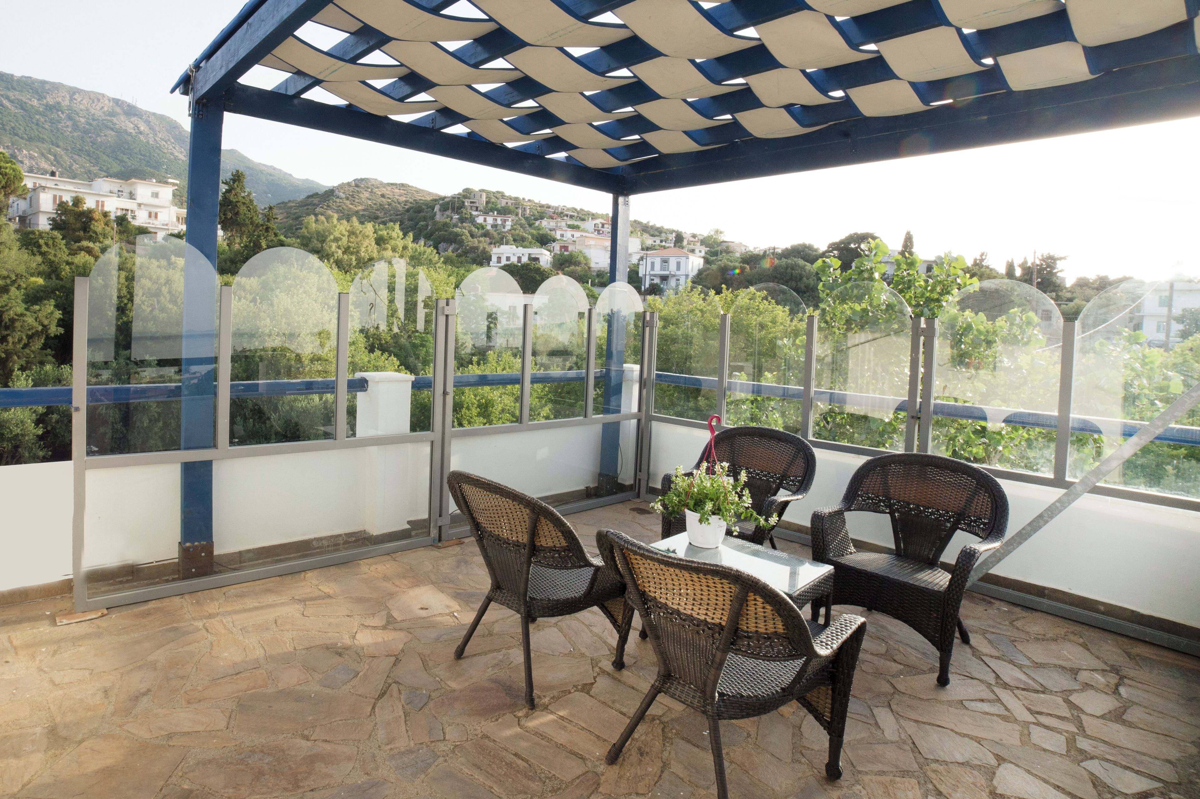 A covered outdoor seating area with wicker chairs and a table, overlooking a scenic hillside with buildings.