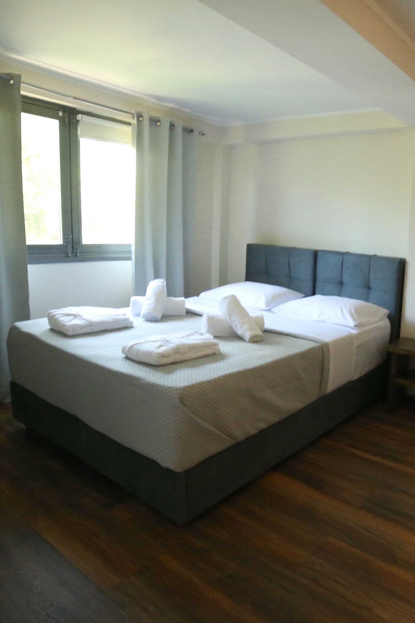 Una cama bien hecha con sábanas y toallas blancas, un cabecero oscuro y una ventana con cortinas.