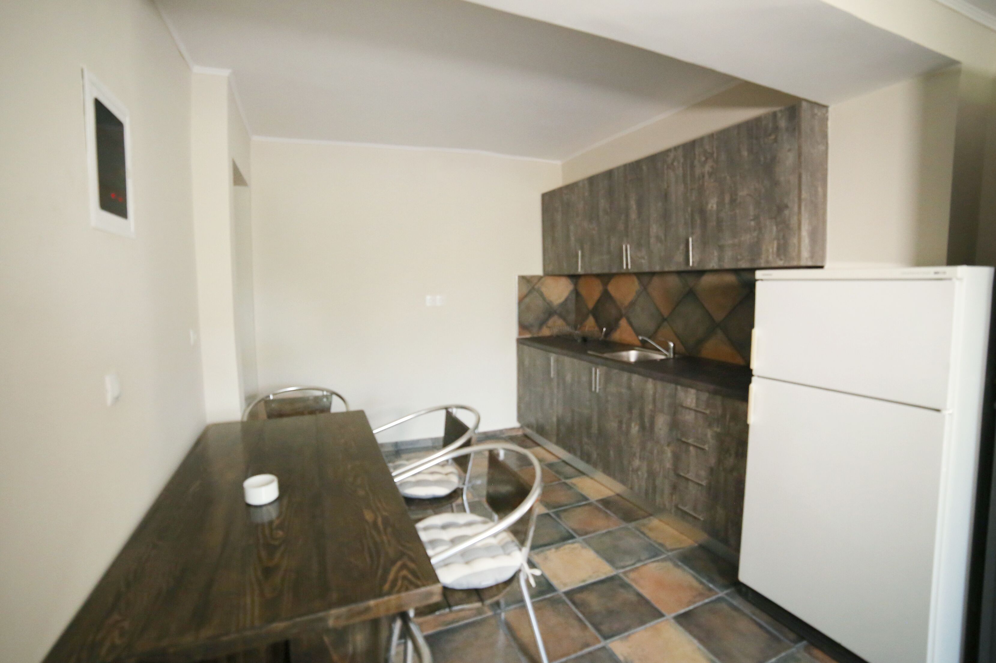 A compact kitchen with a wooden dining table, white chairs, a white refrigerator, and a tiled backsplash.