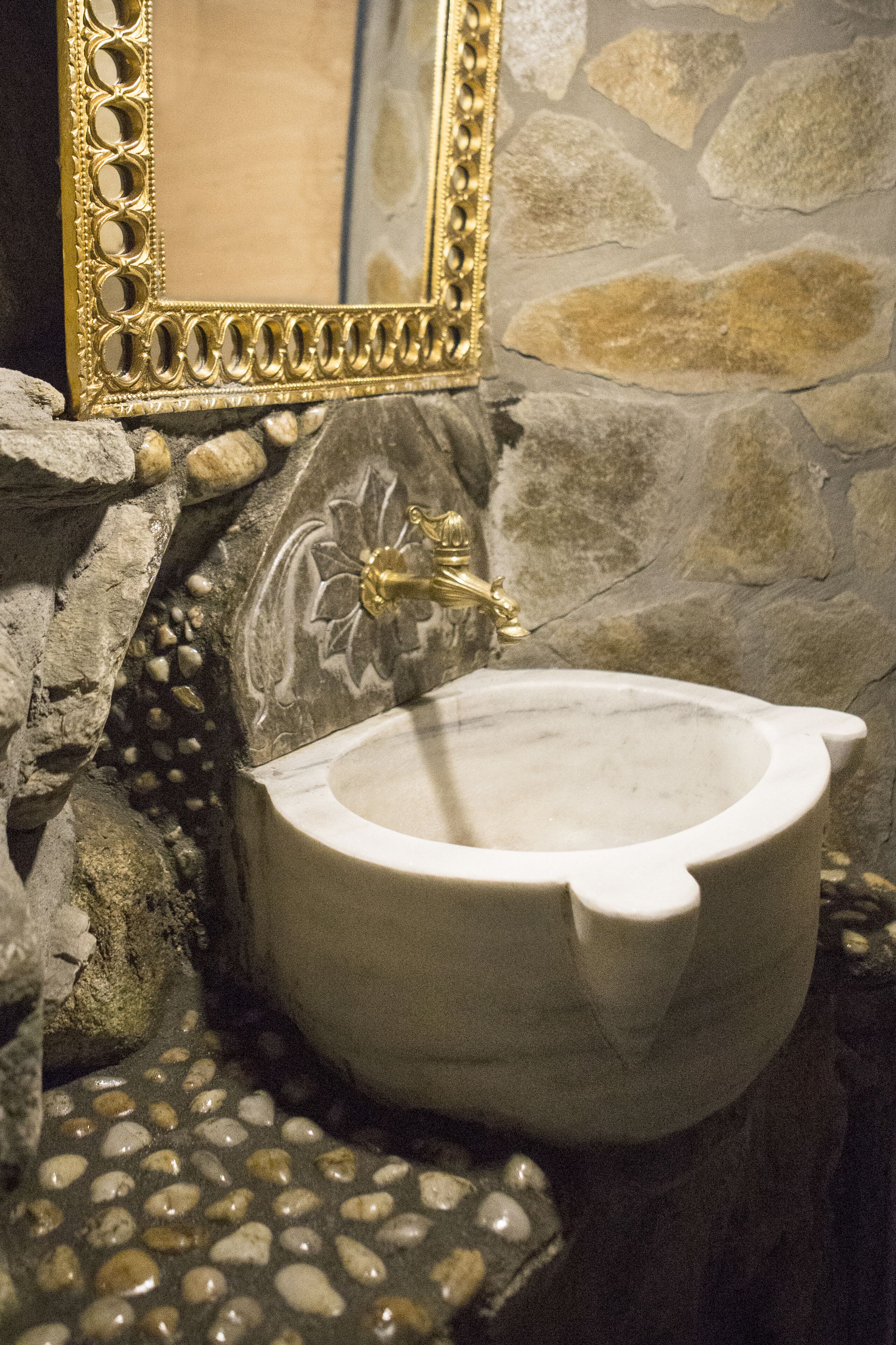 Ein Badezimmer mit Steinwänden, einem мраморный Waschbecken und einem spiegel mit goldener Rahmung.